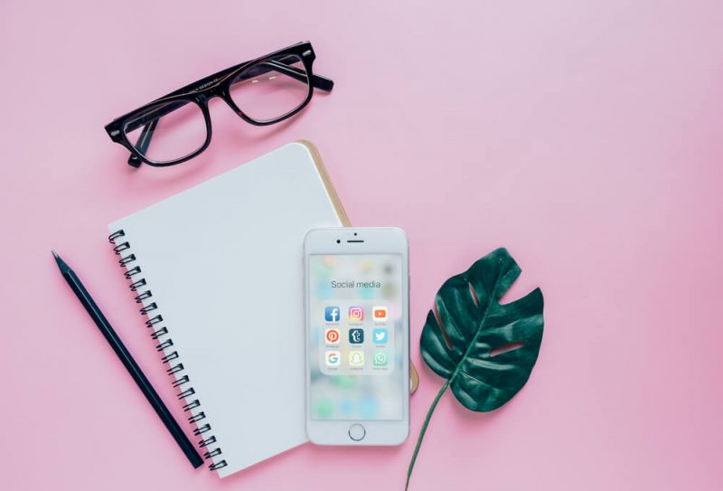 Social Media apps on an iphone with a notepad, pencil, glasses, and a leaf on a pink background